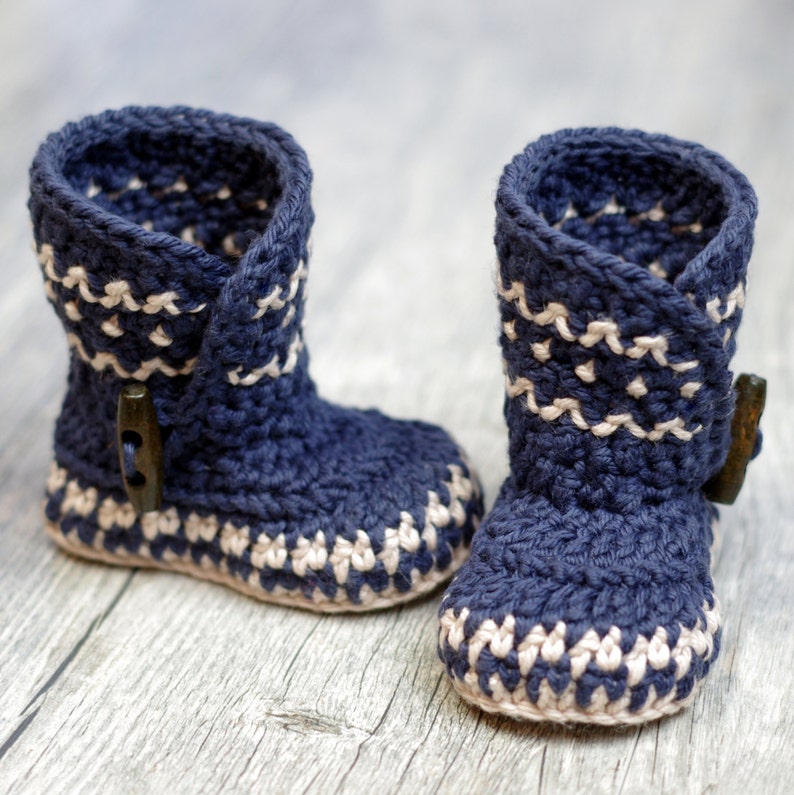 K&ouml;nnte beinhalten: Ein Paar blaue geh&auml;kelte Babyschuhe mit wei&szlig;en Akzenten. Die Schuhe haben ein Knopfdetail an der Seite.