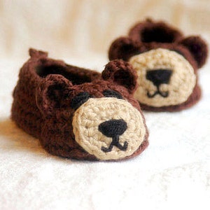 May include: A pair of brown crocheted baby booties with bear faces. The booties have beige snouts and black eyes and noses.
