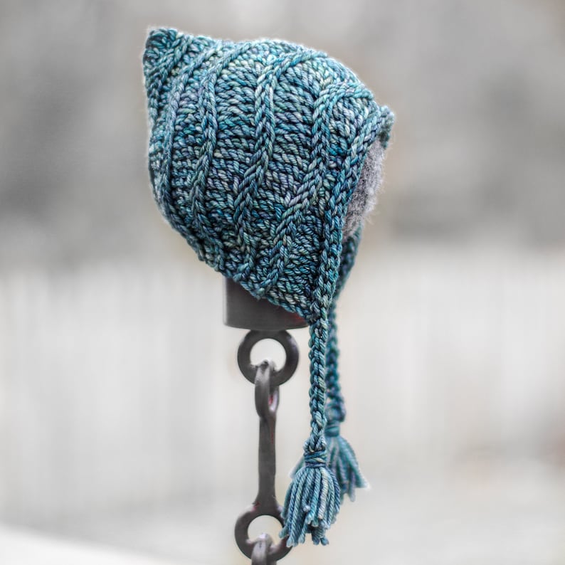 May include: A blue knitted hooded hat with long tassels hanging from the bottom. The hat is displayed on a metal stand.