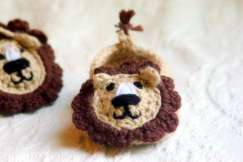 May include: A pair of crocheted baby booties shaped like a lion's head. The booties are brown and beige with a white nose and black eyes.