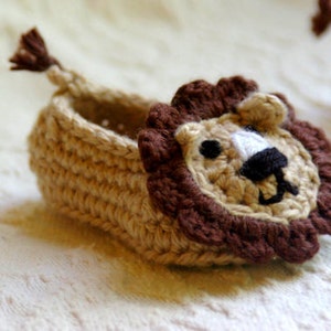 May include: A crocheted baby slipper with a lion face design. The slipper is beige with brown accents and white eyes.