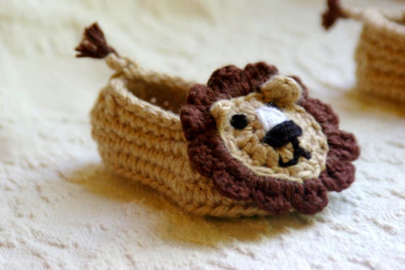 May include: A crocheted baby slipper with a lion face design. The slipper is beige with brown accents and white eyes.