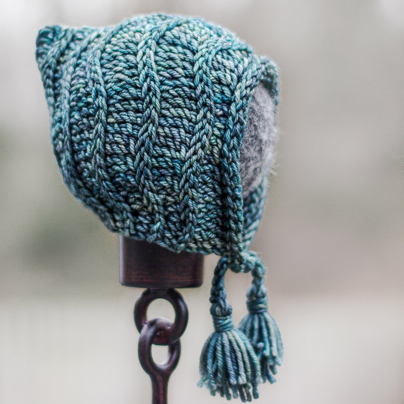 May include: A teal blue knitted hat with a long pointed hood and two tassels. The hat is displayed on a dark brown stand.