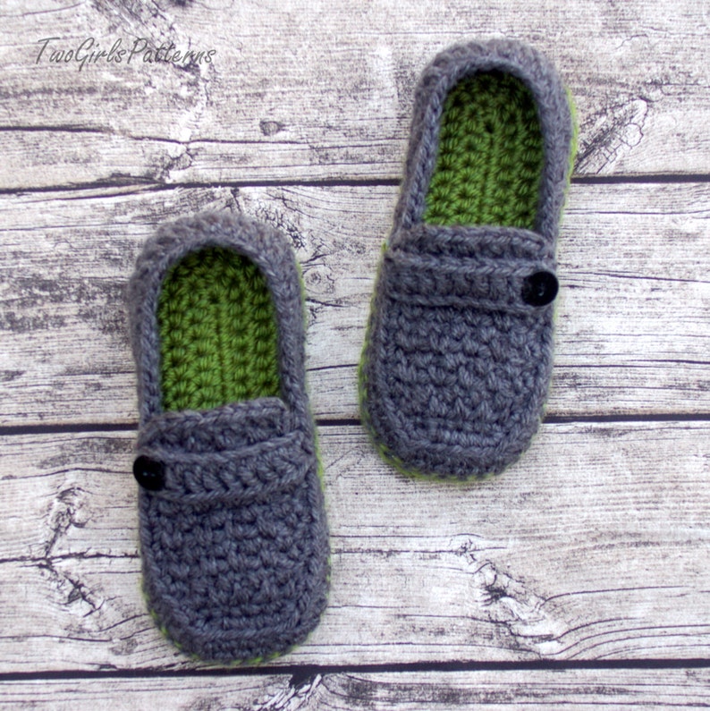 Puede incluir: Un par de zapatillas de crochet grises con forro verde y un bot&oacute;n negro en cada una. Las zapatillas est&aacute;n sobre una superficie de madera.