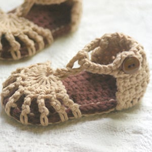 May include: A pair of crocheted baby sandals with brown straps and a brown button. The sandals are made with a beige yarn and have a crocheted design.