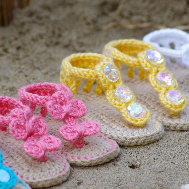 Pu&ograve; includere: Una collezione di sandali in miniatura all'uncinetto con suole beige e bordi rosa o gialli. I sandali rosa presentano decorazioni floreali all'uncinetto, mentre quelli gialli sono impreziositi da gemme in plastica iridescenti e sfaccettate.
