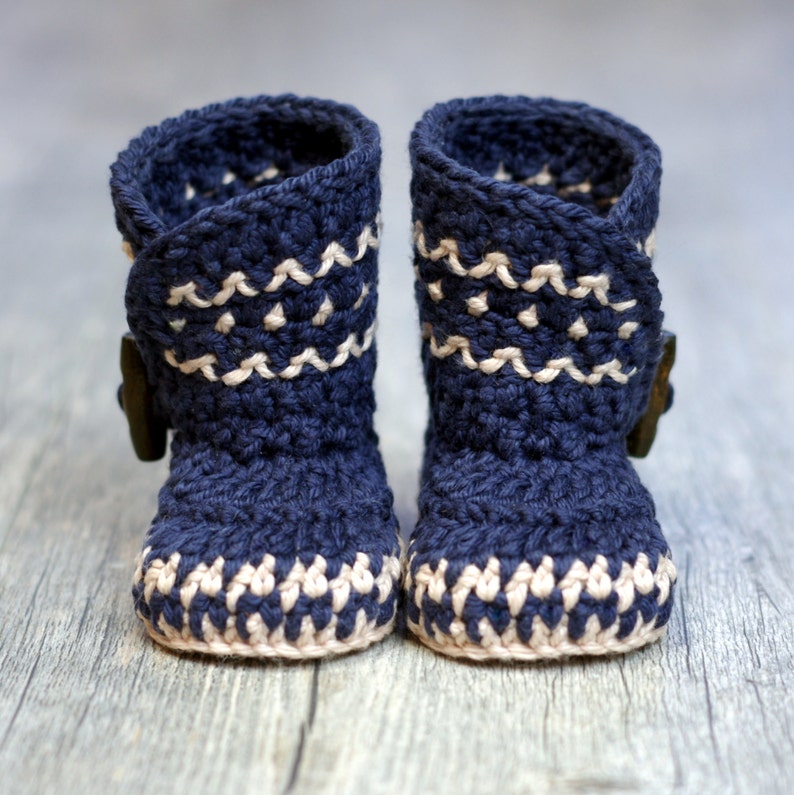 K&ouml;nnte beinhalten: Ein Paar marineblaue geh&auml;kelte Babyschuhe mit wei&szlig;em Besatz und einem Knopfdetail.