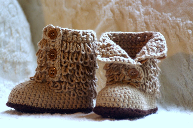 Puede incluir: Un par de botines de beb&eacute; de ganchillo beige con ribetes marrones y botones marrones.