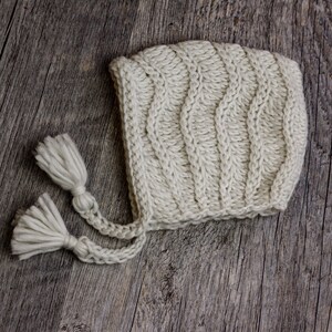 May include: A white knitted baby bonnet with two long tassels. The bonnet is made of a soft, chunky yarn and has a simple, classic design.