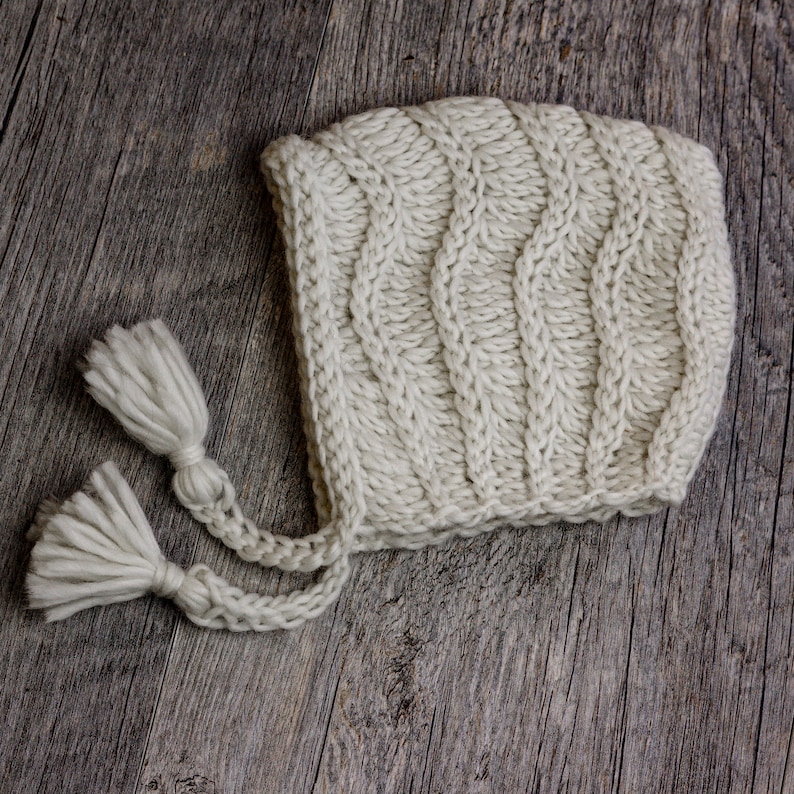 May include: A white knitted baby bonnet with two long tassels. The bonnet is made of a soft, chunky yarn and has a simple, classic design.