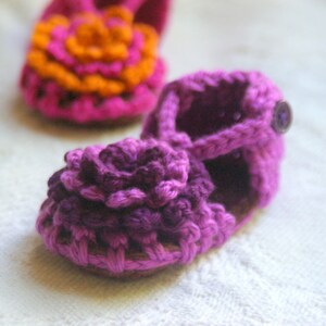 May include: A pair of crocheted baby shoes. One shoe is pink with an orange flower and the other is purple with a purple flower. Both shoes have a button on the strap.