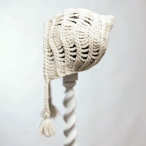 May include: A crocheted baby bonnet with a long tassel. The bonnet is made of a cream-colored yarn and has a delicate, openwork design.