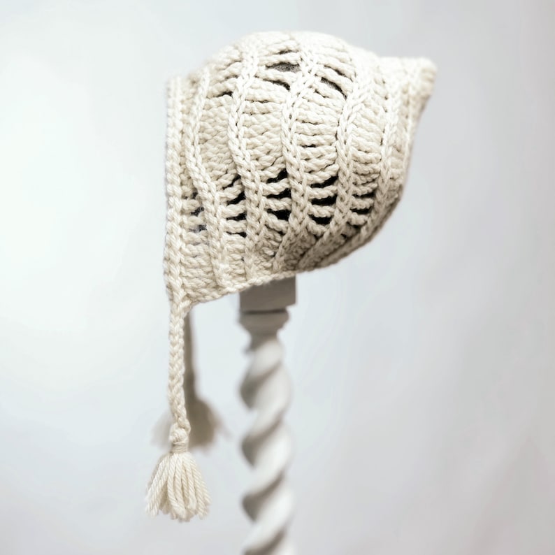 May include: A crocheted baby bonnet with a long tassel. The bonnet is made of a cream-colored yarn and has a delicate, openwork design.
