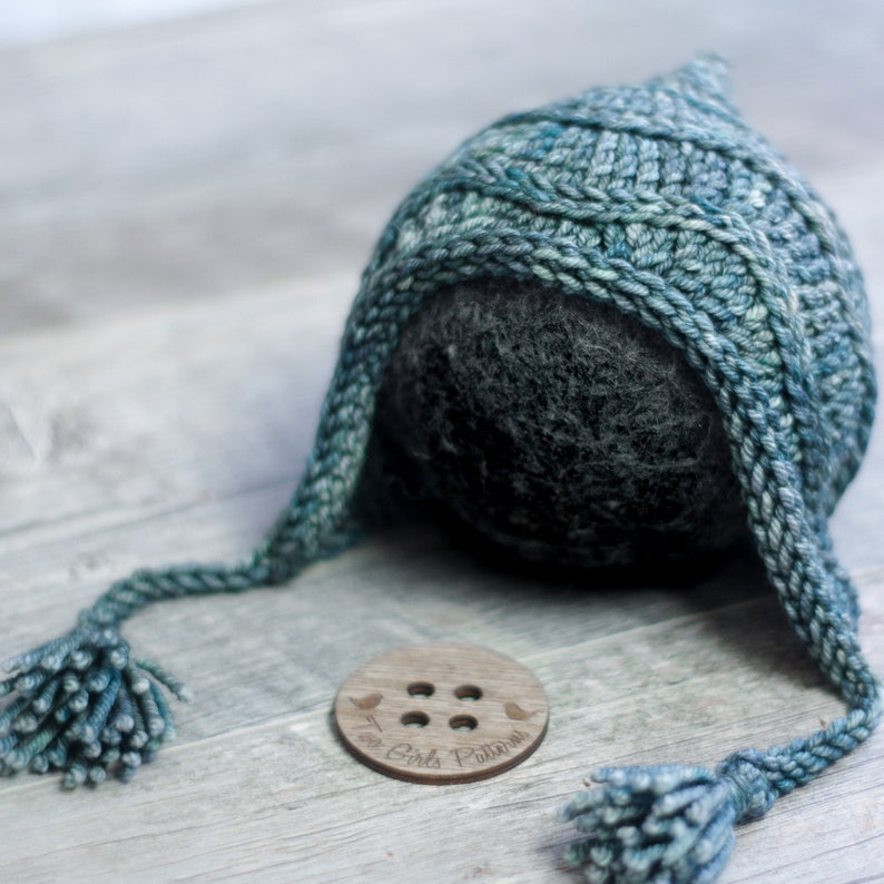 May include: A teal blue knitted baby hat with long ties and tassels. The hat is lying on a wooden surface with a wooden button that says "The Girls Pattern".