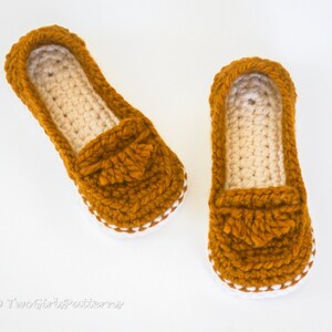 May include: A pair of crocheted slippers in a mustard yellow color with a white trim. The slippers have a loafer style with a crocheted tassel detail.