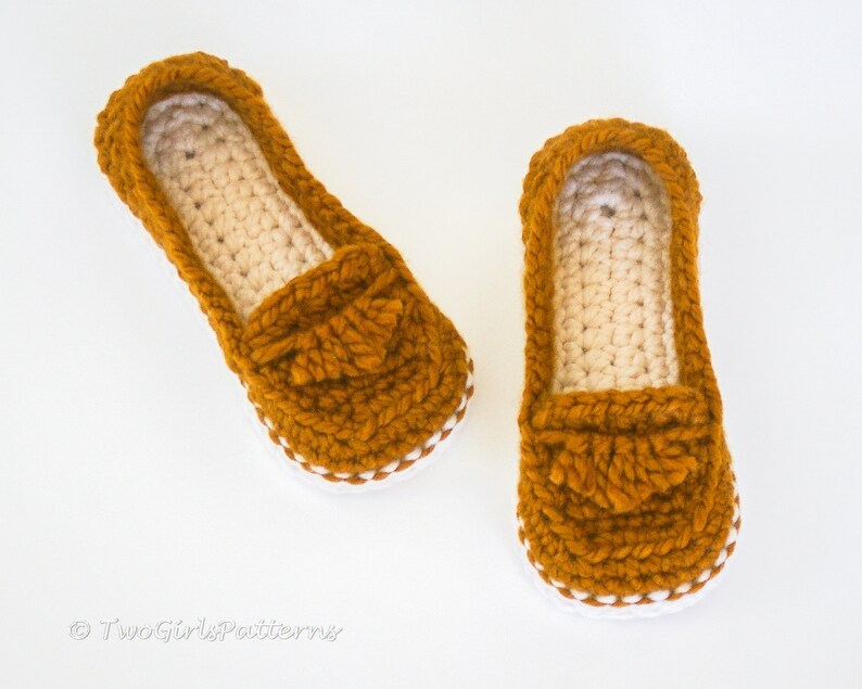 May include: A pair of crocheted slippers in a mustard yellow color with a white trim. The slippers have a loafer style with a crocheted tassel detail.