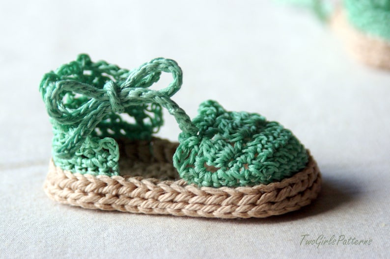 May include: A pair of crocheted baby sandals with a green upper and a beige sole. The sandals have a bow on the top.