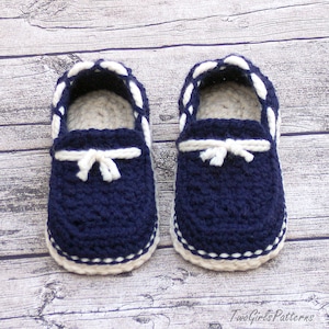 Puede incluir: Un par de zapatos de beb&eacute; de ganchillo azul marino con ribetes blancos y un detalle de lazo blanco. Los zapatos est&aacute;n sobre un fondo de madera.