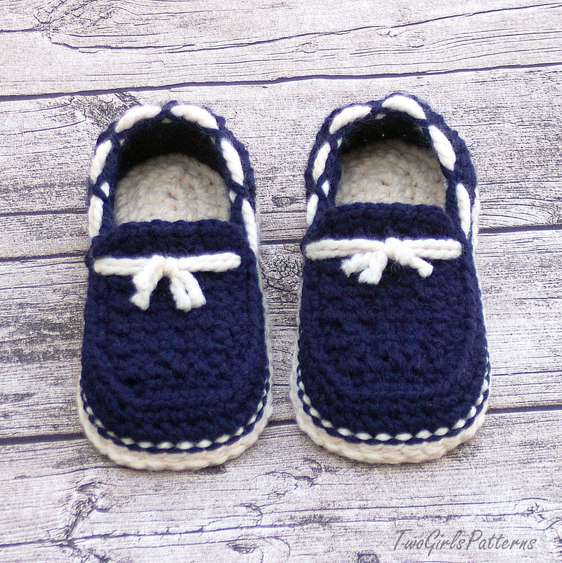 Puede incluir: Un par de zapatos de beb&eacute; de ganchillo azul marino con ribetes blancos y un detalle de lazo blanco. Los zapatos est&aacute;n sobre un fondo de madera.
