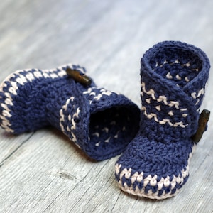 K&ouml;nnte beinhalten: Ein Paar blaue geh&auml;kelte Babyschuhe mit wei&szlig;em Besatz und einem braunen Knopf an jedem Schuh.