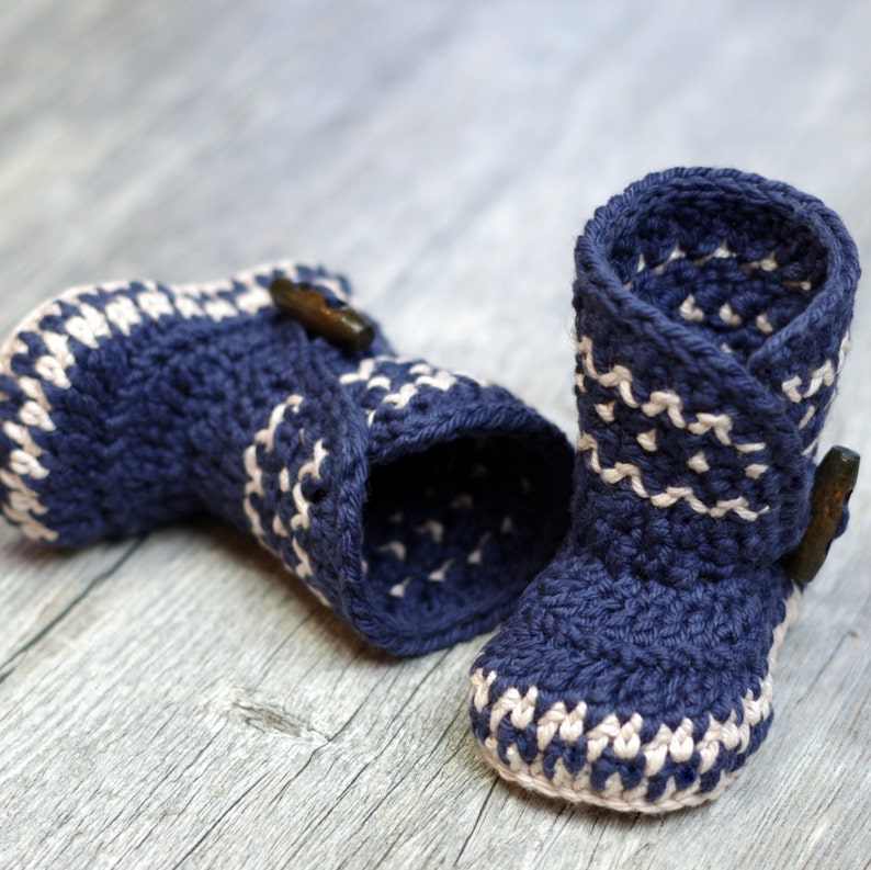 K&ouml;nnte beinhalten: Ein Paar blaue geh&auml;kelte Babyschuhe mit wei&szlig;em Besatz und einem braunen Knopf an jedem Schuh.