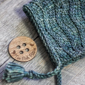 May include: A wooden button with the text "Two Girls Patterns" and two small bird silhouettes. The button is on a wooden surface next to a green knitted hat with a long tassel.