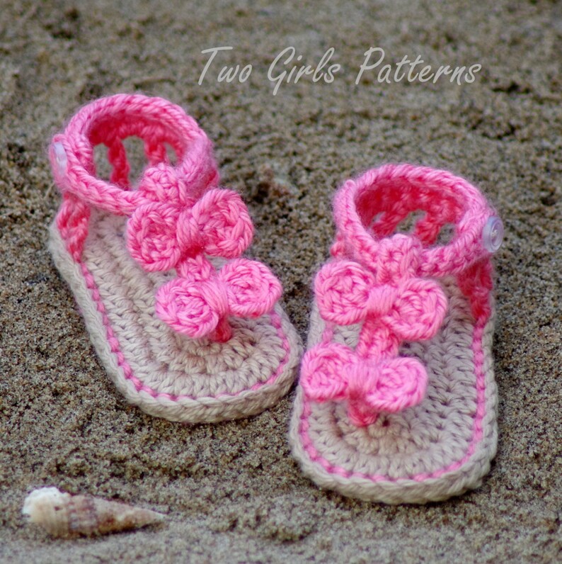 Pu&ograve; includere: Un paio di sandali lavorati all'uncinetto a mano in beige chiaro e rosa acceso, con due fiocchi rosa sovrapposti sul cinturino anteriore e una piccola chiusura a bottone bianco sul cinturino alla caviglia. Il testo Two Girls Patterns &egrave; visibile in alto.