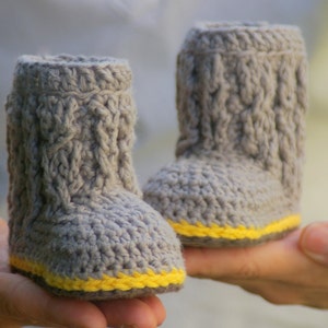 May include: Two gray crocheted booties with yellow trim. The booties are held in the hands of a person.