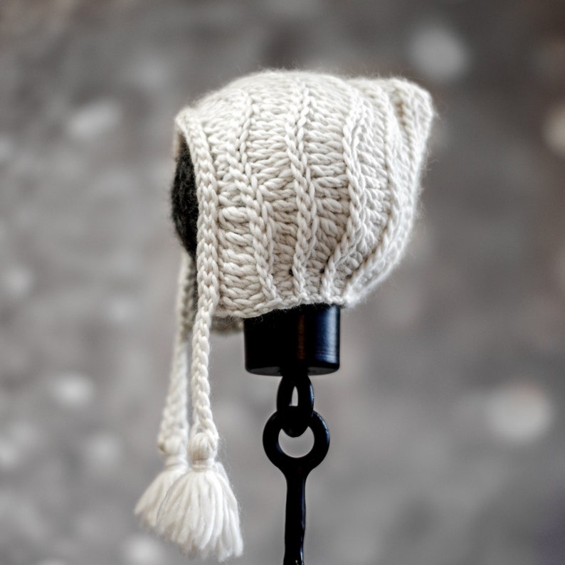 May include: A white crocheted baby bonnet with a long tie and tassels. The bonnet is displayed on a black stand.