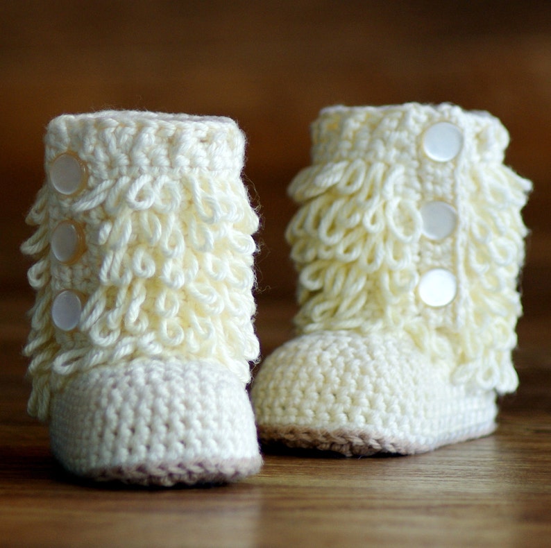 Puede incluir: Un par de botitas de beb&eacute; de crochet blanco con un borde de volantes y botones blancos. Las botitas tienen una suela marr&oacute;n claro.