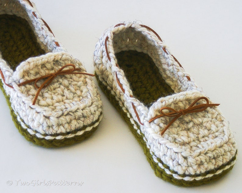 May include: A pair of crocheted slippers with a brown leather strap and bow. The slippers are white with a green trim.
