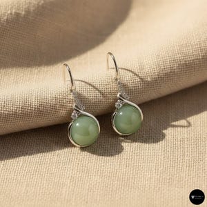 Puede incluir: Un par de pendientes colgantes plateados con una piedra redonda verde claro. Los pendientes tienen un diseño decorativo en espiral con pequeñas piedras de acento transparentes. Los pendientes se exhiben sobre una tela beige.