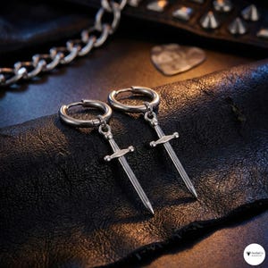 Puede incluir: Un par de pendientes de aro plateados con un colgante de espada. Los pendientes se muestran sobre una superficie negra texturizada, con una cadena plateada y una púa de guitarra en el fondo. Los colgantes de espada tienen empuñaduras y hojas detalladas.