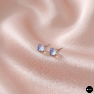 Könnte beinhalten: Ein Paar kleiner, quadratischer Ohrstecker mit Silberstift und Fassung. Die Ohrringe haben einen hellblauen und weißen schillernden Stein. Die Ohrringe werden auf einer rosa Satin-Oberfläche präsentiert.