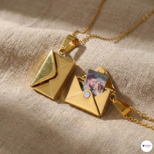 Puede incluir: Un collar con colgante de sobre dorado, abierto para revelar una pequeña fotografía. El colgante está unido a una delicada cadena dorada. El collar se muestra sobre un fondo de tela de color neutro.