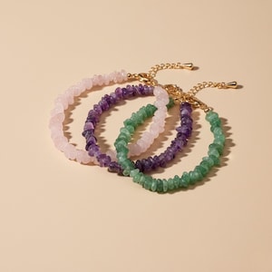 Può includere: Tre braccialetti con pietre preziose rosa, viola e verdi. Ogni braccialetto presenta piccole pietre di forma irregolare infilate insieme. I braccialetti hanno catene e chiusure color oro. Lo sfondo è di colore beige chiaro.