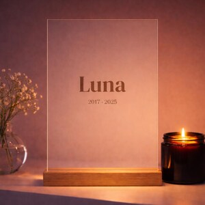 Könnte beinhalten: Ein klarer Acrylschild mit dem eingravierten Namen "Luna" und den Daten "2017-2025", auf einem Holzsockel. Eine brennende Kerze in einem dunklen Glas und eine Vase mit weißen Blumen befinden sich auf beiden Seiten.