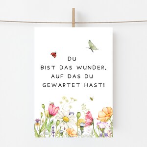 May include: A white print featuring the German text "Du bist das Wunder, auf das du gewartet hast!" with a watercolor floral border. The design includes a ladybug, butterfly, and bee. The flowers are pink, yellow, and white.