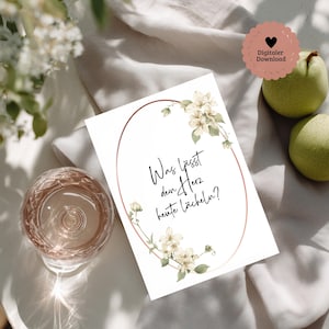 May include: A white card with a rose gold oval frame and floral accents. The card features the text "Was lässt dein Herz heute lächeln?" and is accompanied by a glass of wine and two green pears.