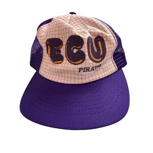 Puede incluir: Una gorra tipo camionero de color morado y blanco con un patrón de cuadrícula en el panel frontal. El frente muestra el texto ECU en letras grandes de color morado oscuro con bordes naranjas y la palabra PIRATES en letras más pequeñas.