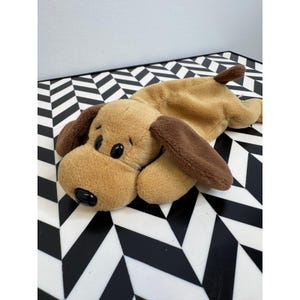 May include: A plush dog toy in shades of brown and tan, with dark brown floppy ears and a small tail. The dog has black button eyes and a black nose. The toy is lying on a black and white geometric patterned surface.