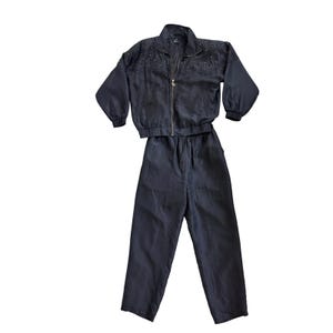 May include: A black two-piece set consisting of a long-sleeve zip-up jacket and matching trousers. The jacket features a high collar, elasticized cuffs, and a decorative yoke adorned with small silver studs. The trousers have a drawstring waistband.