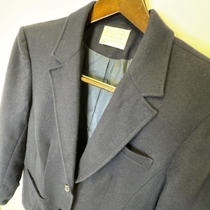 May include: A navy blue blazer with a notched lapel and a single-button closure. The blazer is hanging on a wooden hanger. The interior lining is a darker shade of blue. A label is visible on the inside of the blazer.