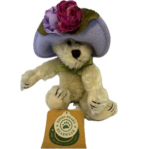 May include: A plush teddy bear wearing a light purple hat adorned with purple and pink flowers. The bear has a green ribbon around its neck and a tag that reads "Boyds Bears Bearwear The Archive Collection."