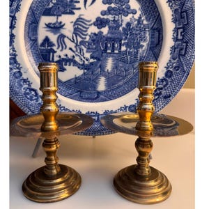 Può includere: Due candelabri in ottone con base rotonda, stelo centrale e piastra circolare per raccogliere la cera. Sullo sfondo un piatto decorativo blu e bianco con un disegno paesaggistico. I candelabri hanno una superficie lucida e riflettente.
