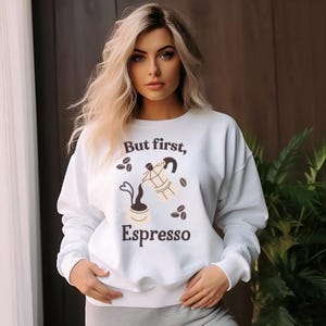 May include: White sweatshirt with the text "But first, Espresso" and coffee-themed graphics. The design includes coffee beans, a coffee pot, and a cup. The sweatshirt is a casual, comfortable style.