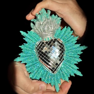 Può includere: Un ornamento decorativo a forma di cuore con un centro a specchio, circondato da raggi glitterati turchesi. La parte superiore presenta un design a fiamma argentata. L'ornamento è tenuto su uno sfondo nero.