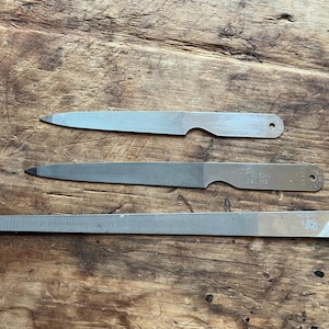 May include: Three silver-colored metal files of varying lengths and shapes. Each file features a textured abrasive surface, a smooth handle section with a small circular hole at the end, and a pointed or rounded tip for precision metalworking tasks.