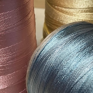 Pode incluir: Três bobinas de linha brilhante e lisa estão dispostas juntas. Uma bobina é rosa antigo, outra é dourada metálica e a terceira é de um azul ardósia frio e cintilante. A linha está enrolada firmemente em torno de cada bobina cilíndrica.