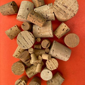 May include: A collection of various cylindrical cork stoppers in different sizes and textures. The natural brown corks feature porous surfaces, some with visible cracks, grain patterns, and markings, scattered across the frame.